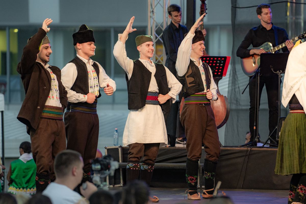 Igrači u narodnoj nošnji podižu ruke na otvorenoj sceni, u pozadini muzičari